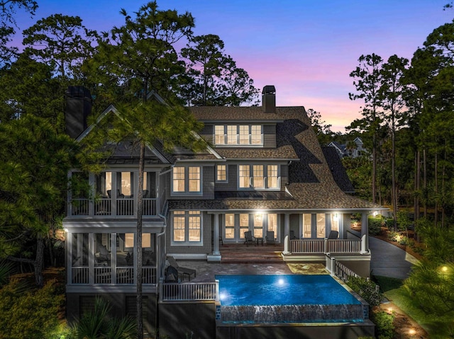 back of house at dusk with roof with shingles, an outdoor pool, a sunroom, and a chimney