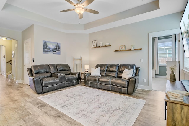 living area featuring a ceiling fan, a tray ceiling, baseboards, light wood finished floors, and arched walkways