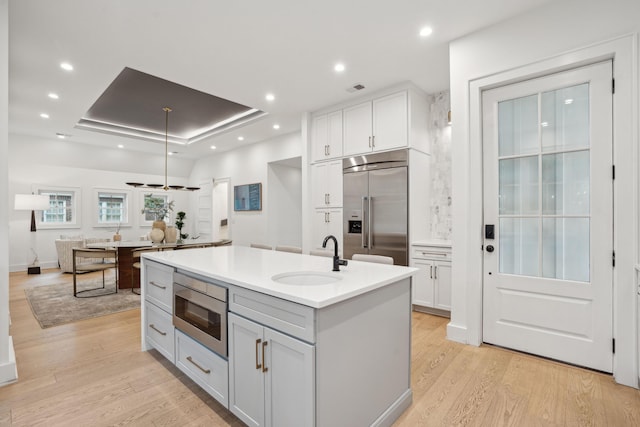 kitchen with a tray ceiling, a center island with sink, a sink, light wood-style floors, and built in appliances