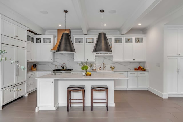 kitchen with a center island with sink, decorative backsplash, premium range hood, white cabinetry, and beam ceiling