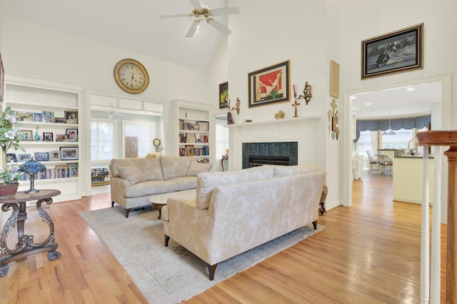 living room featuring high vaulted ceiling, a tile fireplace, light wood-style flooring, a ceiling fan, and built in features