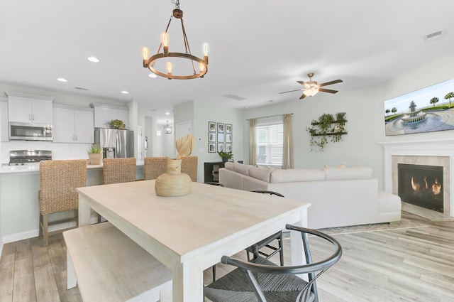 dining area featuring visible vents, light wood-style flooring, recessed lighting, a lit fireplace, and ceiling fan with notable chandelier