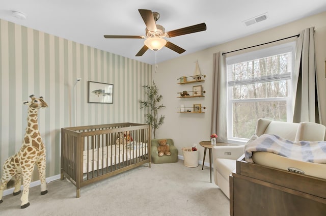 carpeted bedroom featuring visible vents, a ceiling fan, a nursery area, wallpapered walls, and baseboards