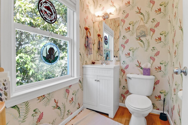 half bath with toilet, wood finished floors, wallpapered walls, baseboards, and vanity