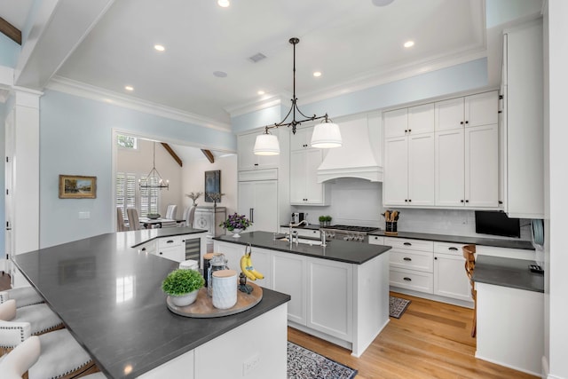 kitchen with custom exhaust hood, white cabinetry, an island with sink, hanging light fixtures, and a chandelier