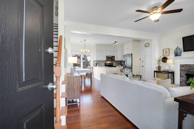 living room featuring ornamental molding, a brick fireplace, dark wood-style flooring, and ceiling fan with notable chandelier