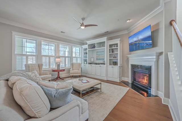 living area featuring baseboards, a fireplace with flush hearth, ceiling fan, wood finished floors, and crown molding