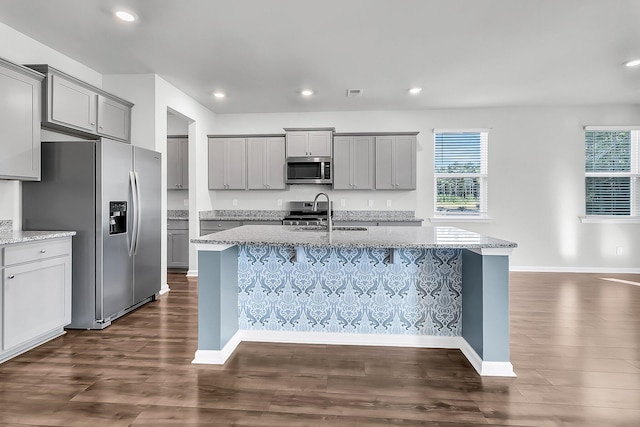 kitchen with dark wood-type flooring, an island with sink, sink, light stone countertops, and appliances with stainless steel finishes