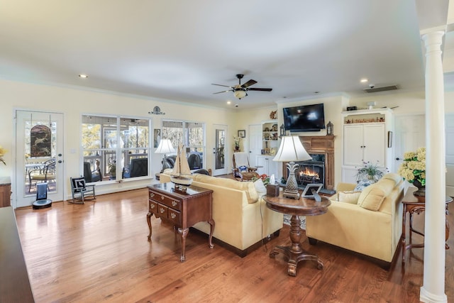 living area featuring a warm lit fireplace, decorative columns, ceiling fan, ornamental molding, and wood finished floors