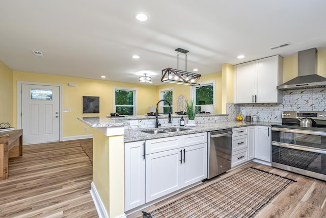 kitchen featuring visible vents, a sink, appliances with stainless steel finishes, a peninsula, and wall chimney range hood