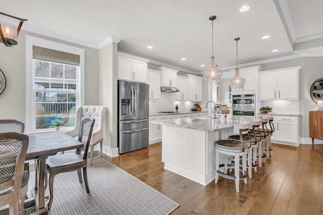 kitchen with light stone counters, appliances with stainless steel finishes, pendant lighting, a kitchen island with sink, and white cabinets