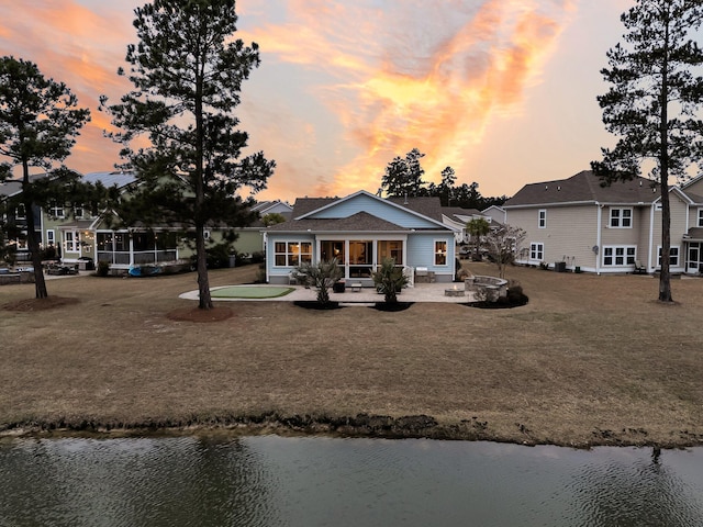 back house at dusk with a water view, a patio area, and a lawn