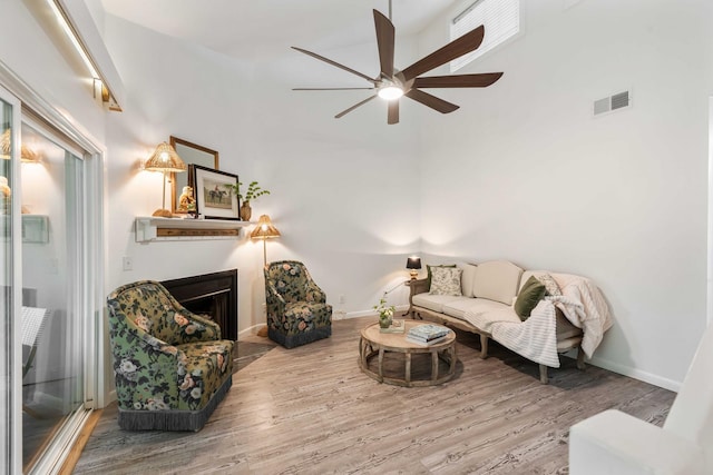 living room featuring wood finished floors, a ceiling fan, a towering ceiling, a fireplace, and baseboards
