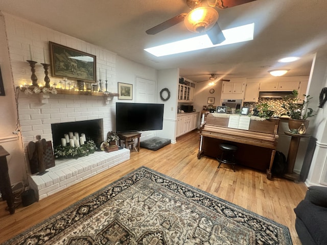 living area with ceiling fan, light wood-style flooring, a brick fireplace, and a skylight