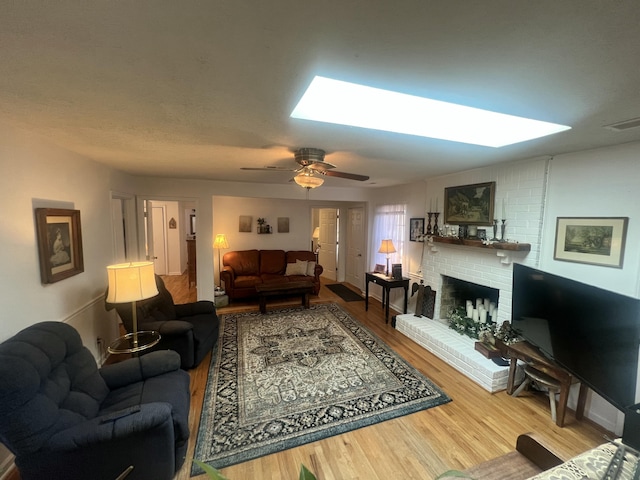 living area with a ceiling fan, wood finished floors, visible vents, a skylight, and a brick fireplace