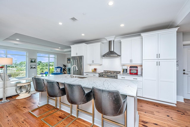 kitchen with a tray ceiling, high end fridge, wall chimney exhaust hood, and an island with sink