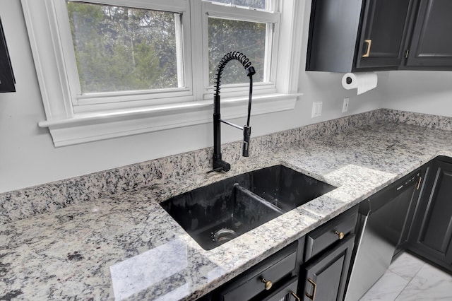 kitchen with light stone counters, a sink, marble finish floor, dark cabinetry, and stainless steel dishwasher