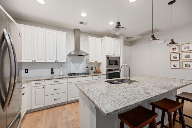 kitchen featuring wall chimney exhaust hood, an island with sink, appliances with stainless steel finishes, and hanging light fixtures