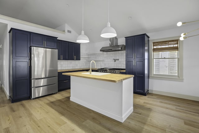 kitchen with wood counters, wall chimney exhaust hood, decorative backsplash, an island with sink, and stainless steel appliances