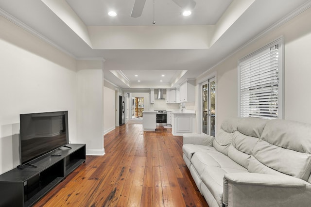 living area with a tray ceiling, wood-type flooring, ornamental molding, ceiling fan, and baseboards