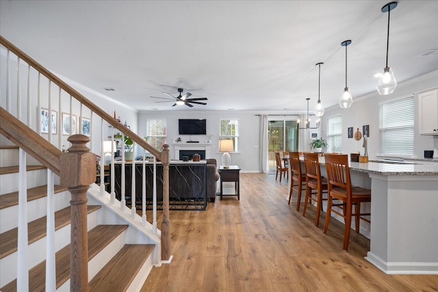 living room featuring light wood-style flooring, ornamental molding, ceiling fan with notable chandelier, a fireplace, and stairs