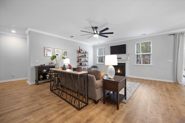 living room with a ceiling fan, baseboards, light wood-style flooring, a warm lit fireplace, and ornamental molding