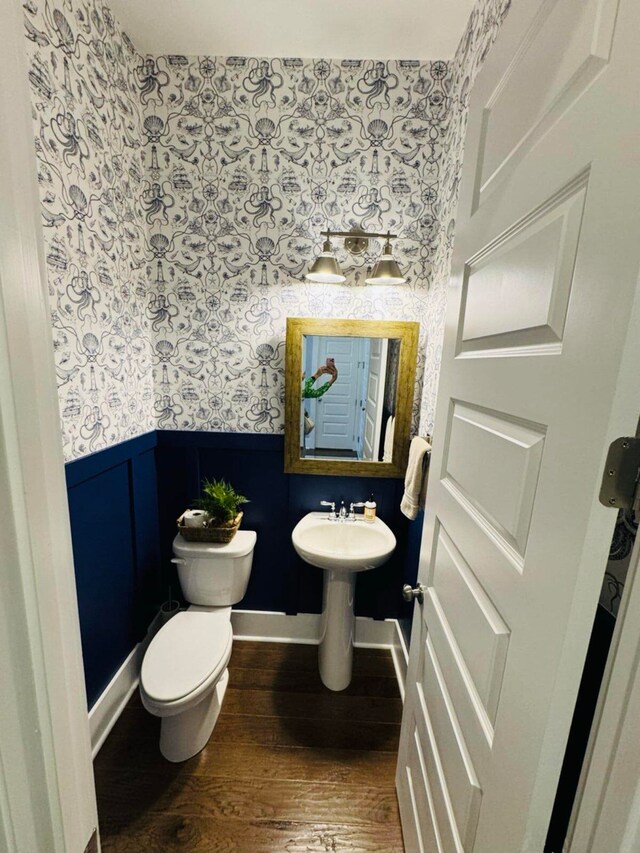 half bath featuring wood finished floors, baseboards, wallpapered walls, wainscoting, and toilet