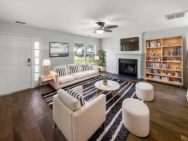 living area featuring a glass covered fireplace, dark wood-style flooring, a ceiling fan, and baseboards