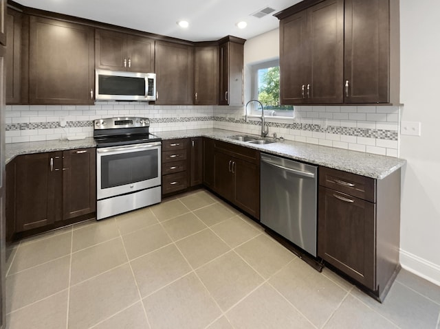 kitchen featuring appliances with stainless steel finishes, a sink, dark brown cabinets, light stone counters, and decorative backsplash