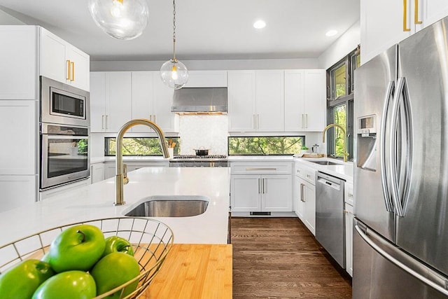 kitchen featuring wall chimney range hood, sink, white cabinetry, hanging light fixtures, and stainless steel appliances