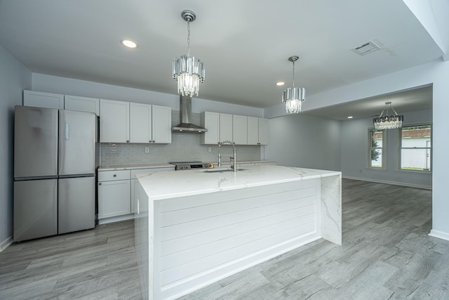 kitchen with light wood-type flooring, white cabinetry, stainless steel fridge, wall chimney exhaust hood, and a center island with sink