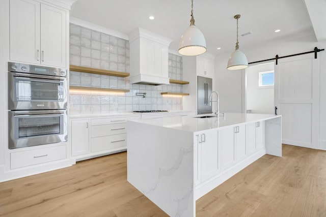 kitchen featuring white cabinetry, hanging light fixtures, a center island with sink, appliances with stainless steel finishes, and a barn door