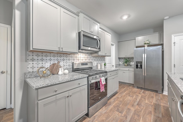 kitchen with stainless steel appliances, wood finished floors, gray cabinets, decorative backsplash, and light stone countertops