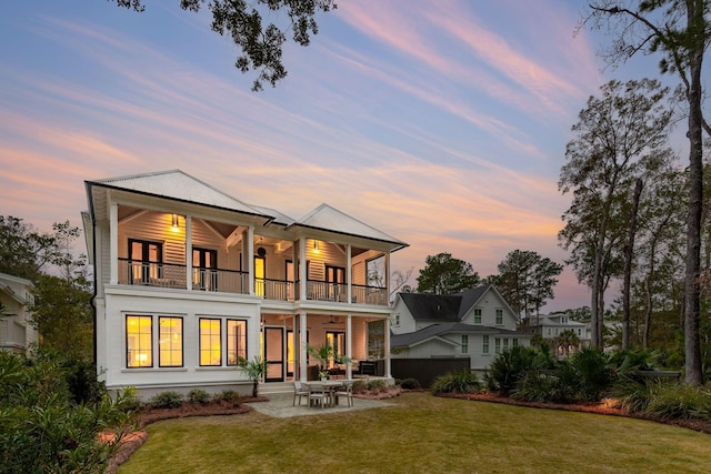 back house at dusk featuring a balcony, a patio area, ceiling fan, and a yard