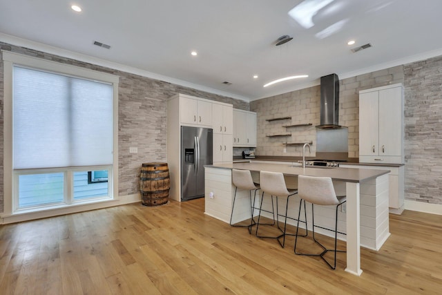 kitchen with stainless steel fridge, a center island with sink, white cabinetry, and wall chimney range hood