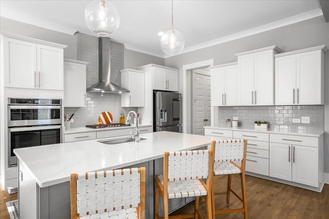 kitchen featuring wall chimney range hood, an island with sink, stainless steel appliances, pendant lighting, and dark wood-type flooring