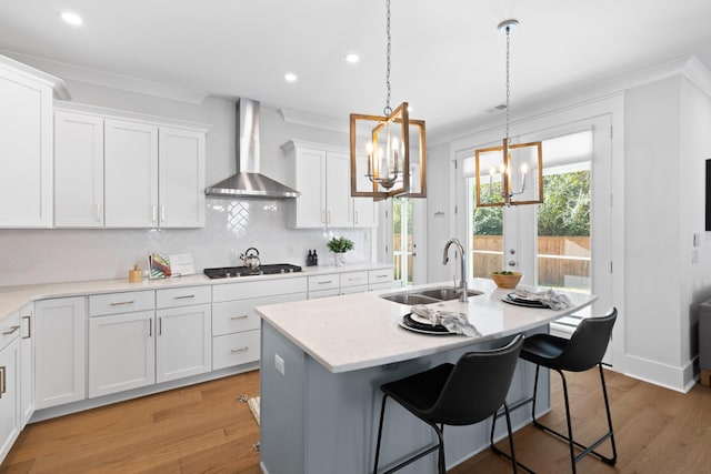 kitchen featuring a kitchen island with sink, wall chimney range hood, sink, white cabinetry, and stainless steel gas cooktop