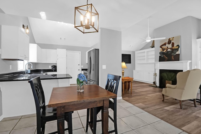 dining space featuring built in features, a fireplace, ceiling fan with notable chandelier, light tile patterned floors, and vaulted ceiling