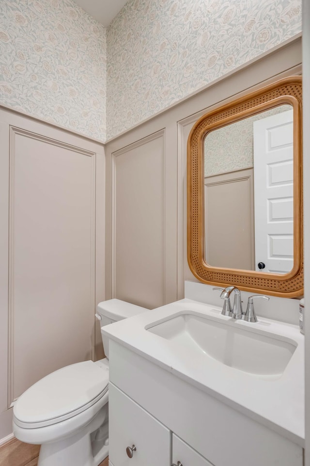 half bath with a decorative wall, toilet, wallpapered walls, and vanity