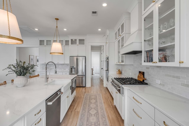 kitchen featuring custom range hood, appliances with stainless steel finishes, light wood-style floors, hanging light fixtures, and a sink