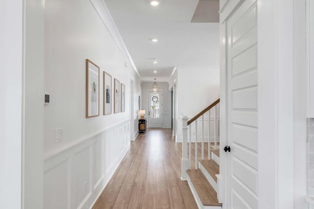 corridor featuring stairs, wainscoting, crown molding, a decorative wall, and light wood-type flooring