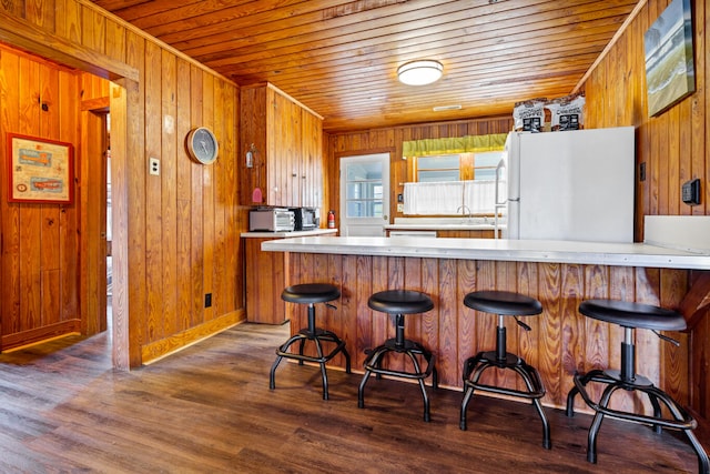 kitchen with dark wood-type flooring, white refrigerator, kitchen peninsula, wood walls, and a kitchen bar