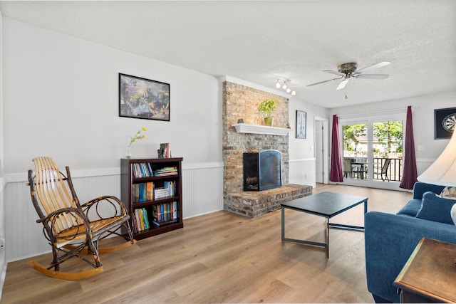 living area featuring a ceiling fan, a textured ceiling, a brick fireplace, wood finished floors, and a wainscoted wall