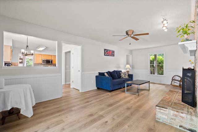 living area with light wood-style floors, a textured ceiling, ceiling fan with notable chandelier, and a wainscoted wall