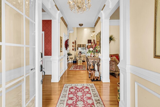 entrance foyer featuring a chandelier, light wood finished floors, wainscoting, wallpapered walls, and ornate columns