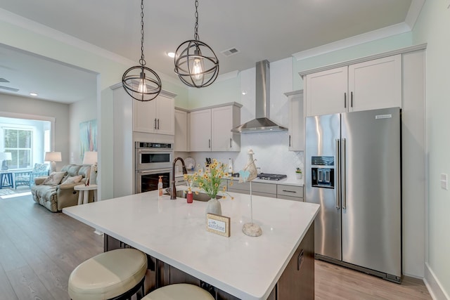 kitchen with a breakfast bar, appliances with stainless steel finishes, an island with sink, wall chimney range hood, and backsplash