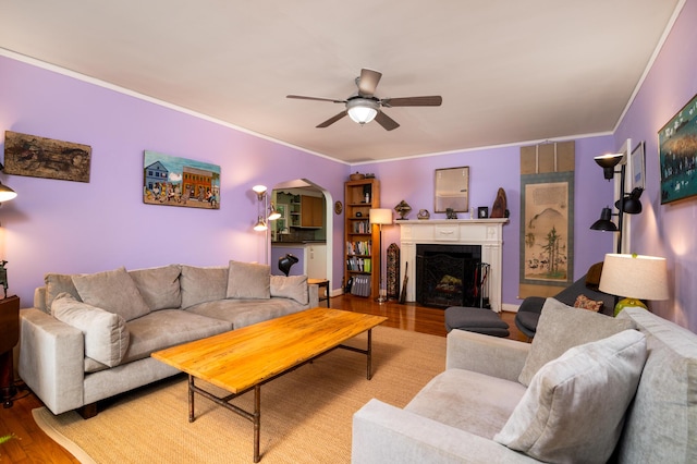 living area with wood finished floors, a ceiling fan, arched walkways, ornamental molding, and a fireplace