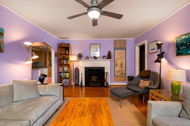 living area featuring wood finished floors, arched walkways, crown molding, ceiling fan, and a fireplace with flush hearth