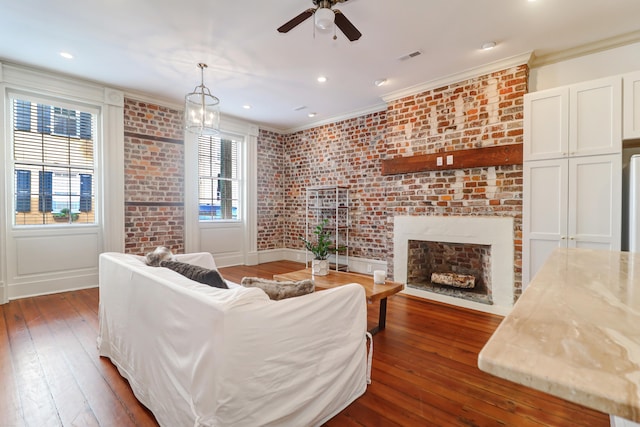 living room featuring ceiling fan with notable chandelier, ornamental molding, a brick fireplace, hardwood / wood-style flooring, and brick wall