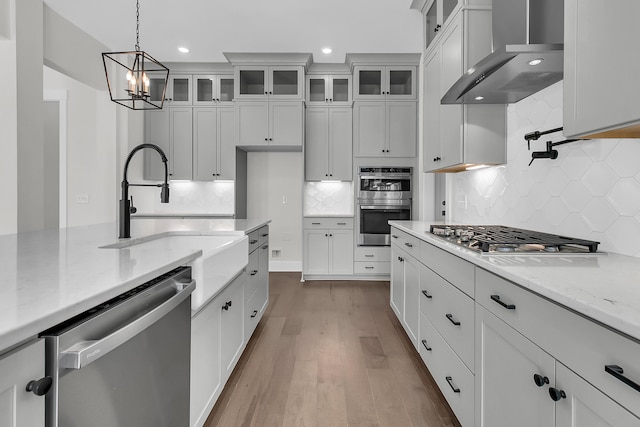 kitchen with pendant lighting, wall chimney range hood, appliances with stainless steel finishes, light stone countertops, and white cabinets